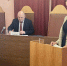 На изображении представлен зал заседаний, где проходит официальное мероприятие. В центре внимания — женщина, стоящая за трибуной и, вероятно, выступающая с речью. Она одета в пиджак в клетку и черную блузку. На заднем плане виден мужчина в темном костюме. - Юргамышский муниципальный округ